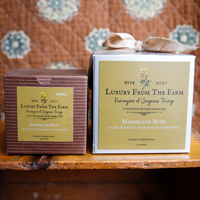 Two boxes of 'Luxury From The Farm' on a wooden surface with a patterned fabric background. marshland moss scent. candle and vessel in ne and refill in the other box